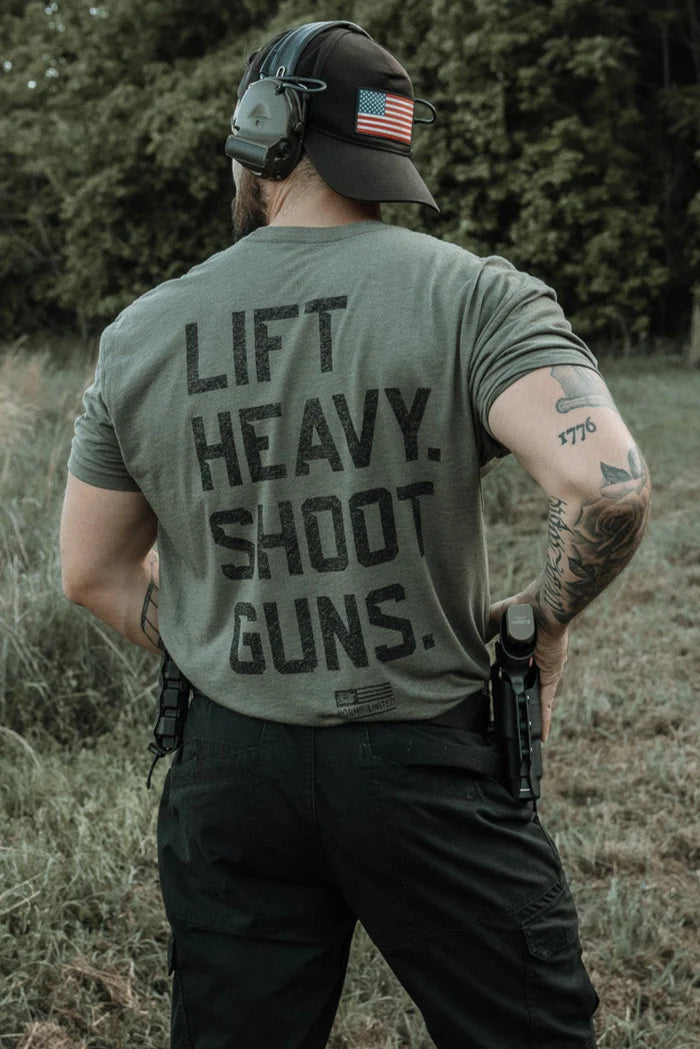 A man stands outdoors in a Tango Charlie Apparel "Lift Heavy. Shoot Guns." shirt, wearing headphones and an American flag cap, with tattoos and pistols on his hips; trees and grass form the background.