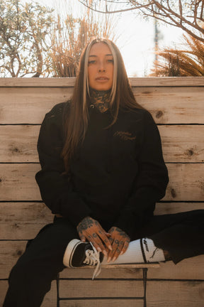 A woman with long brown hair and tattoos sits outdoors on a chair, exuding resilience in the Relentless Betrayal Vengeance Hoodie, black pants, and black and white sneakers, one leg propped up against a wooden fence backdrop with plants.