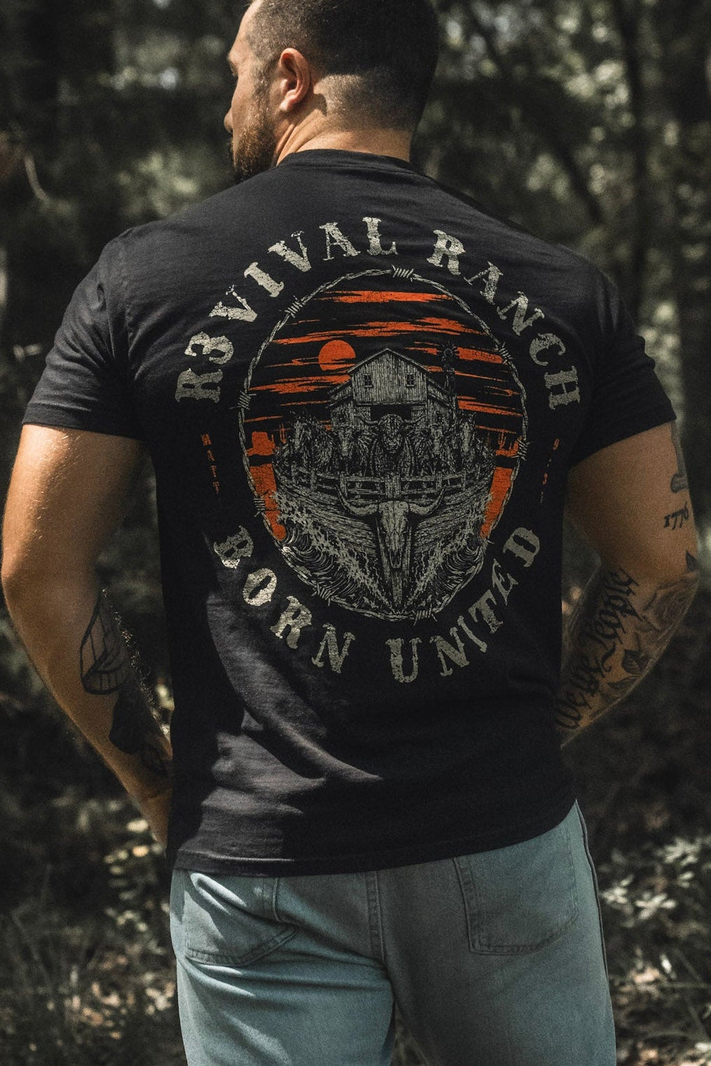 A man stands outdoors with his back to the camera, wearing a black Revival Ranch Premium Tee from Born United Private Label—a piece of Christian apparel—and light blue jeans.