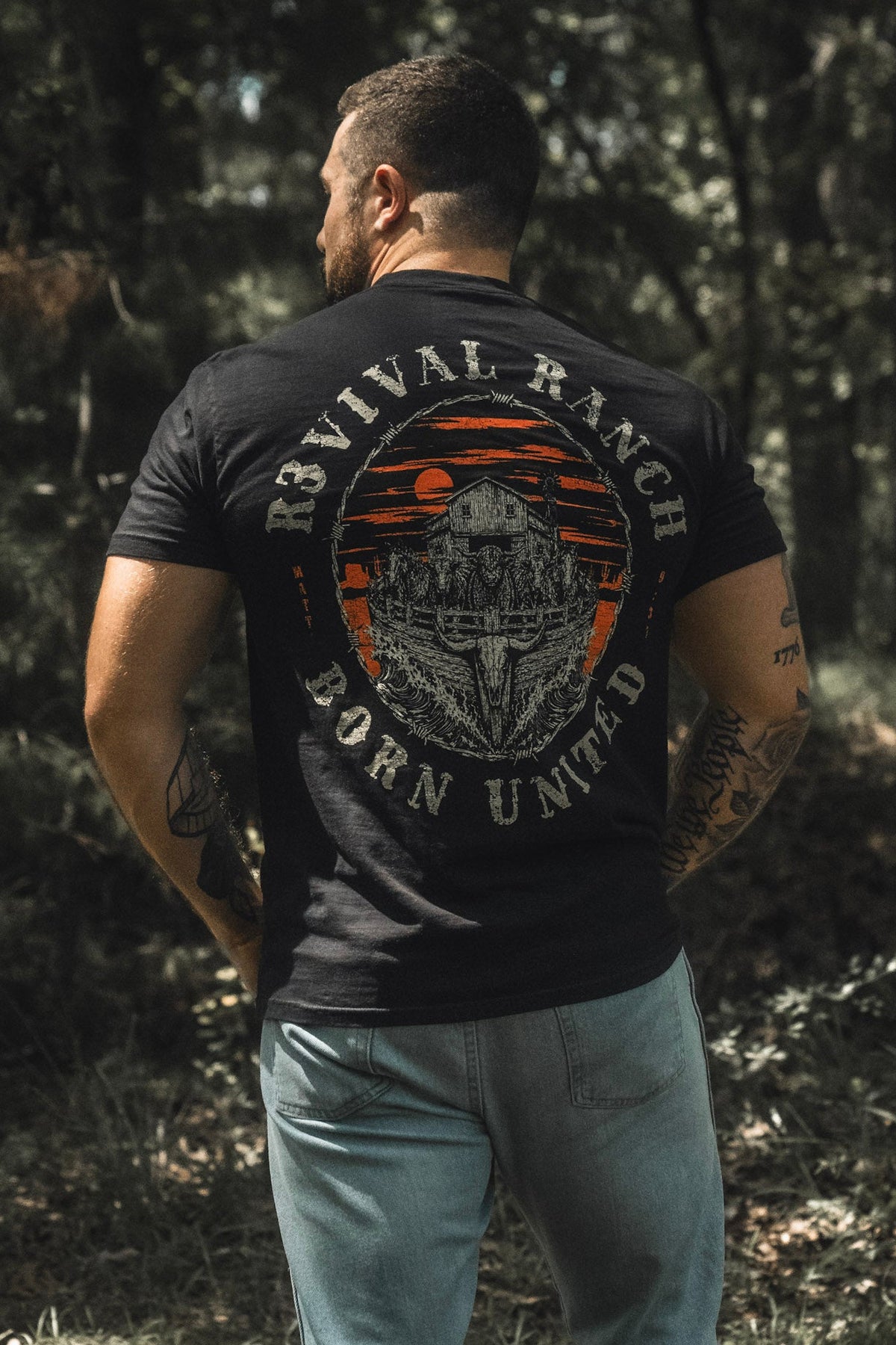 A man stands outdoors with his back to the camera, wearing a black Revival Ranch Premium Tee from Born United Private Label—a piece of Christian apparel—and light blue jeans.