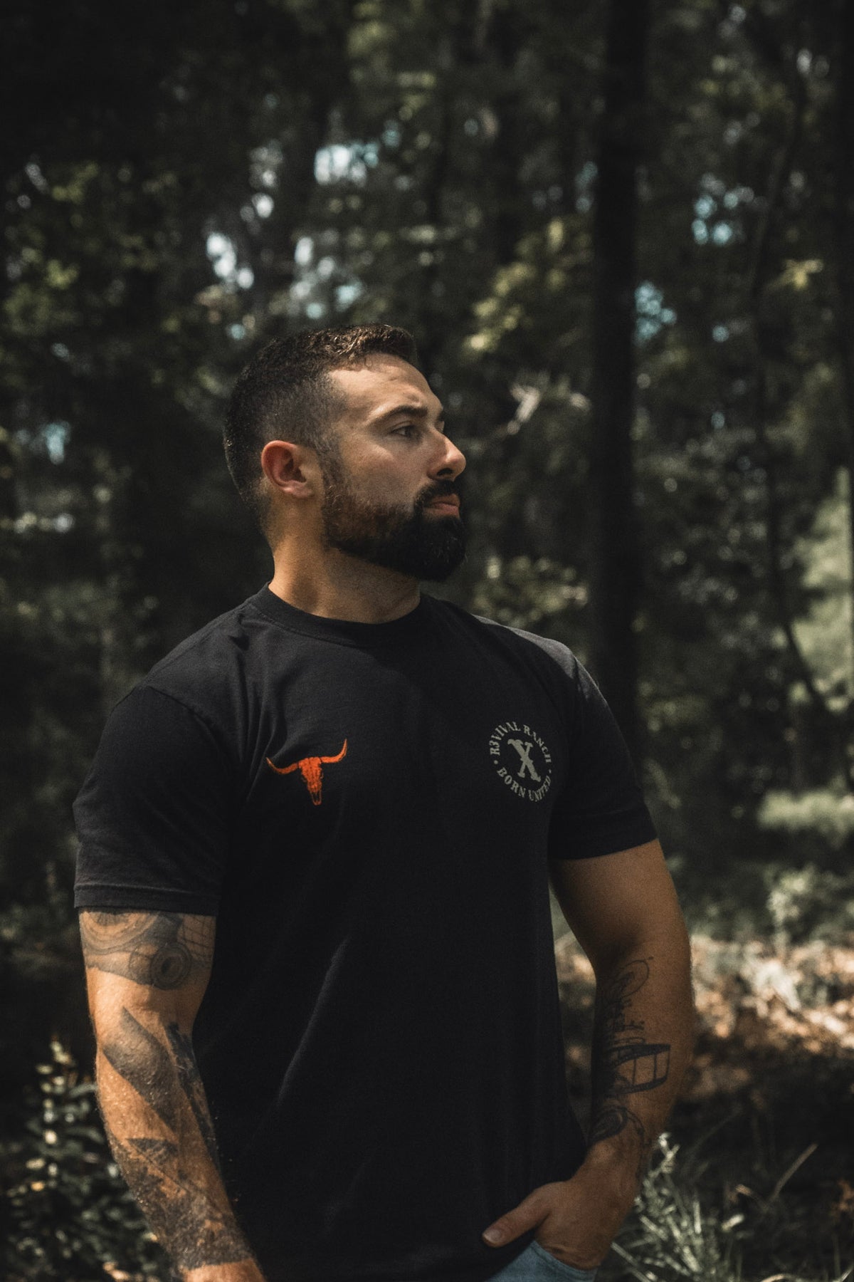 A bearded, tattooed man stands outdoors in a shaded forest area, wearing a black Revival Ranch Premium Tee by Born United Private Label featuring front graphics, and looking to the side.