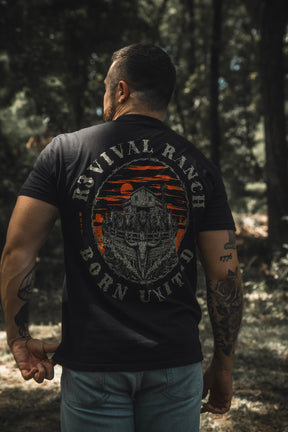 A man with tattoos, wearing jeans and a black Revival Ranch Premium Tee from Born United Private Label, stands outdoors in a wooded area, facing away from the camera.