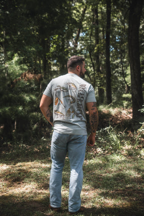 A man with tattoos stands outdoors in a wooded area, facing away from the camera, wearing the Mucker Bucket Bait DNA Premium Tee and light blue jeans.