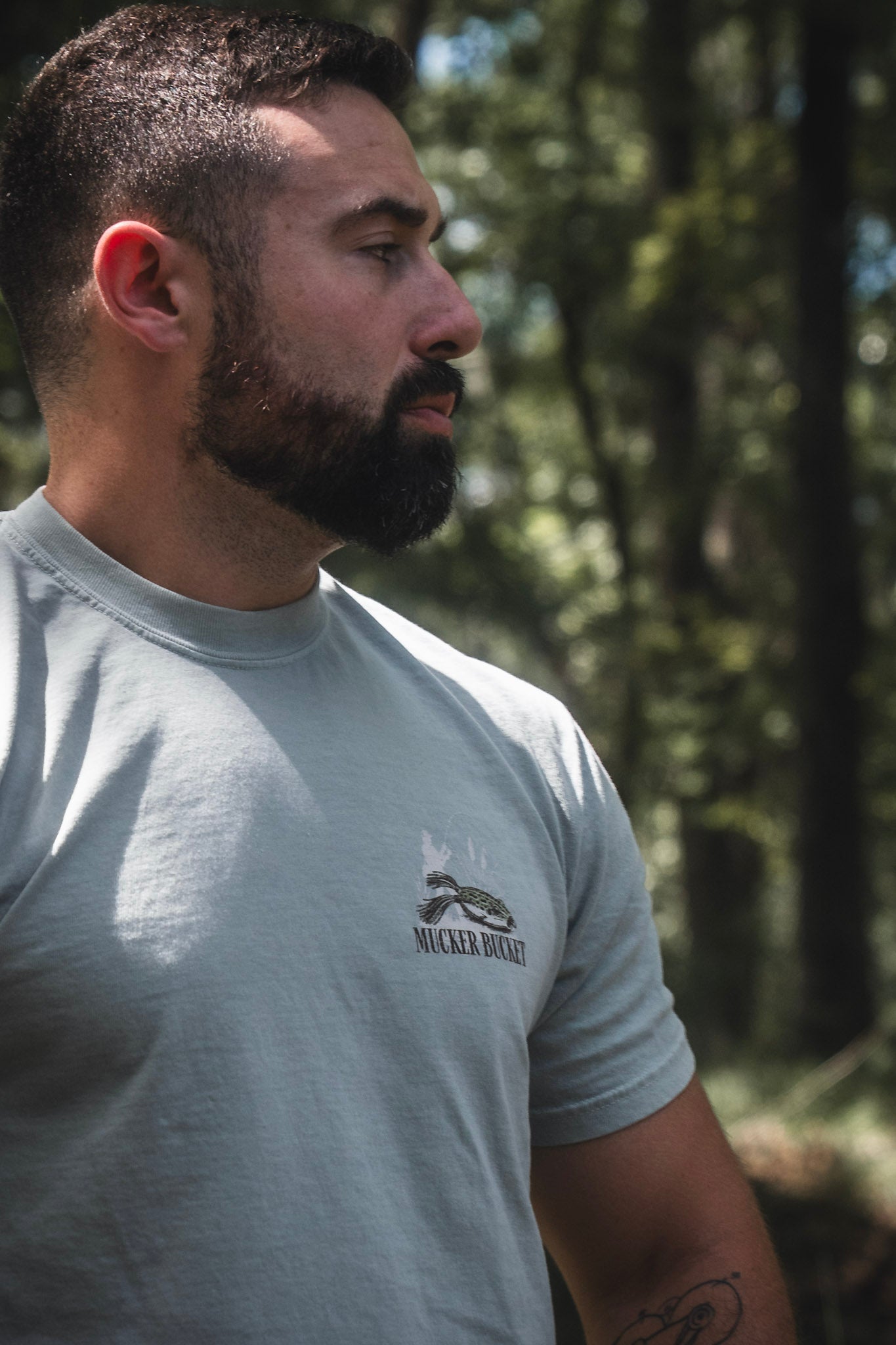 A man with a beard stands outdoors in a pale Mucker Bucket Bait DNA Premium Tee, looking off to the side. Crafted from soft ring-spun cotton, this premium relaxed fit tee by Mucker Bucket complements the natural backdrop of blurred trees and greenery.