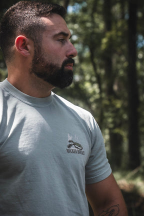 A man with a beard stands outdoors in a pale Mucker Bucket Bait DNA Premium Tee, looking off to the side. Crafted from soft ring-spun cotton, this premium relaxed fit tee by Mucker Bucket complements the natural backdrop of blurred trees and greenery.