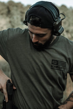 A bearded man in a backward cap and ear protection wears a Tango Charlie Apparel "Lift Heavy. Shoot Guns." shirt outdoors, looking down as he reaches for a handgun at his waist.