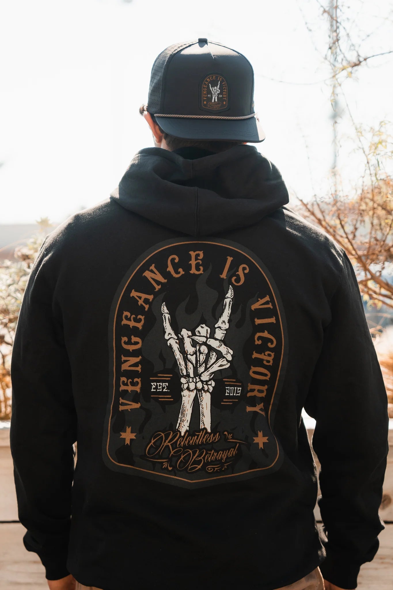 A person wears the Relentless Betrayal Vengeance Hoodie—black with a matching cap. The hoodie’s back features an illustrated skeleton with swords and the words “Vengeance is Victory,” symbolizing resilience and empowerment.