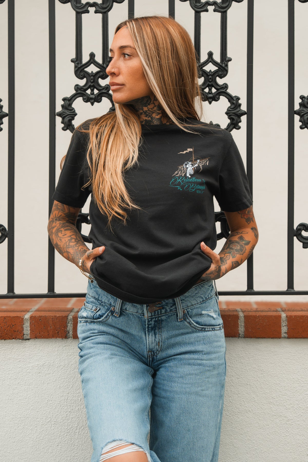 A person with long hair and tattoos stands against an ornate iron fence, wearing the Relentless Betrayal Glorious Redemption Premium Tee crafted from high-quality jersey cotton, paired with blue jeans, hands in pockets, gazing to the side.