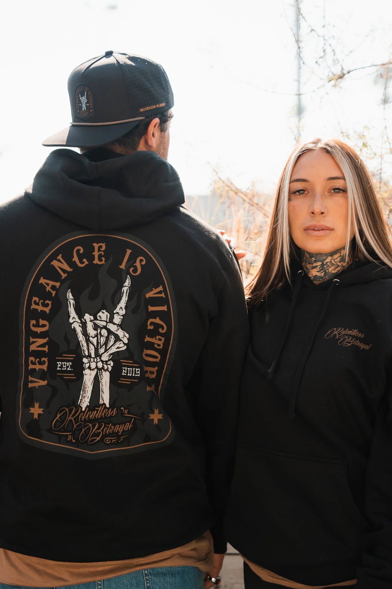 A man and woman wear black Relentless Betrayal Vengeance Hoodies; his shows a skeletal hand gripping a sword with "Vengeance Is Victory," while hers displays "Relentless Betrayal" on the front.