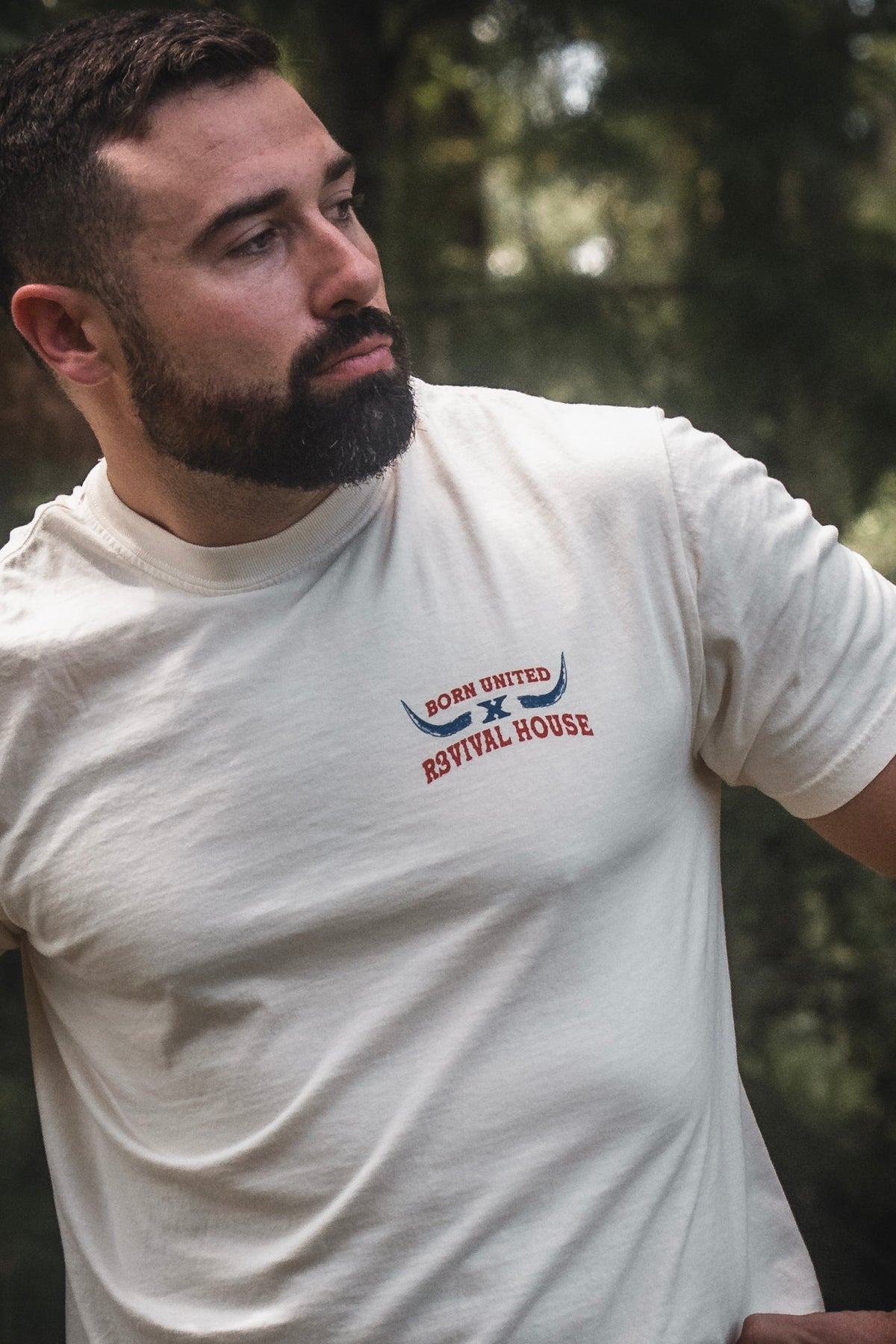 A bearded man in a white Revival House Premium Tee by Born United Private Label stands outdoors, looking to the side, embodying the spirit of a faith-based ranch and showcasing Christian apparel inspired by R3VIVAL Ranch.