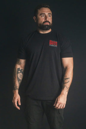 A muscular man with a beard stands against a dark background, wearing the Born United Private Label “We Remember: 25th Anniversary” tee and pants, his tattooed arms on display—a powerful image of honor and resilience.