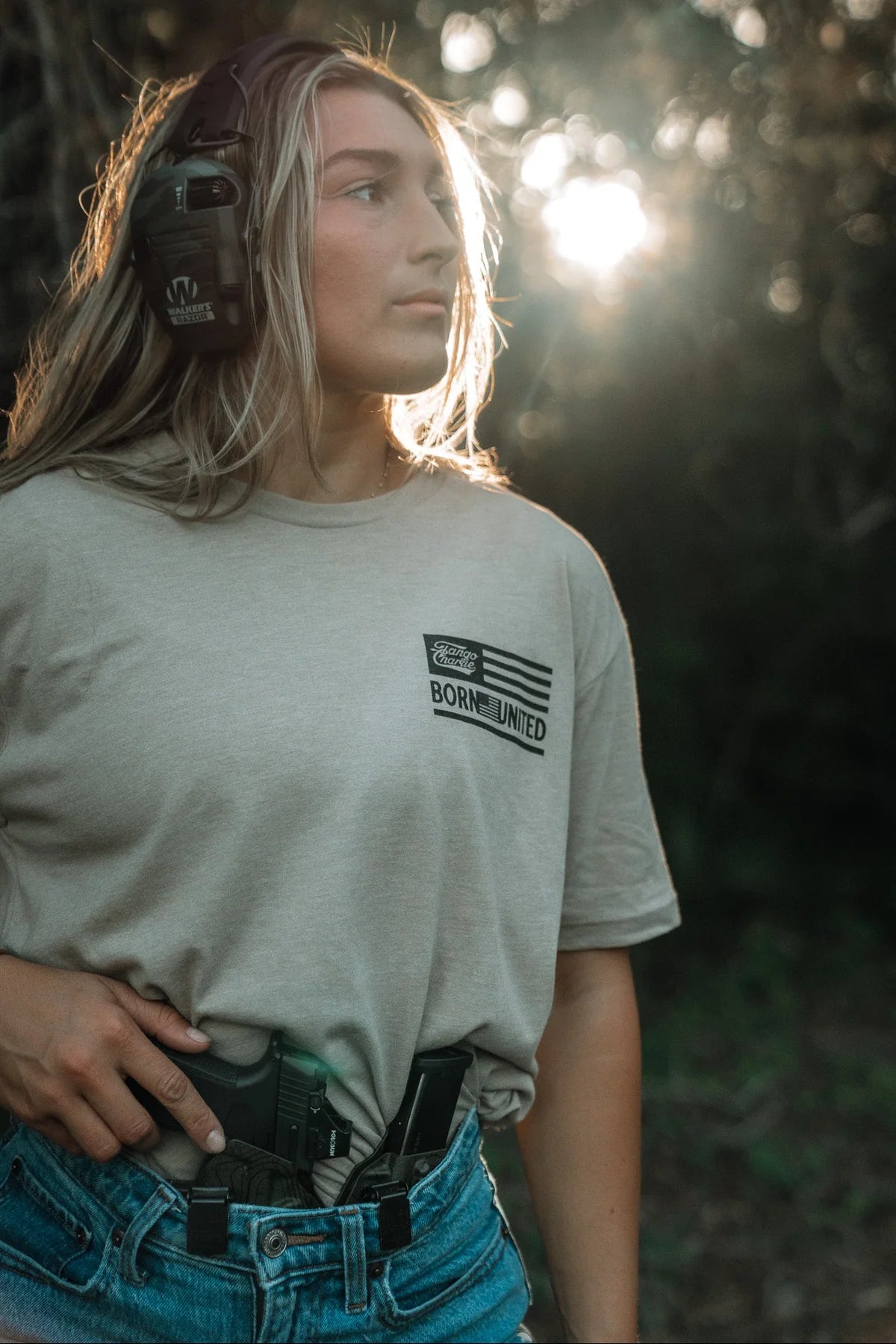 A woman wearing earmuffs and the LHSG - Tan Tee by Tango Charlie Apparel stands outdoors in sunlight, a holstered handgun tucked into her jeans, looking off to the side with a focused expression.