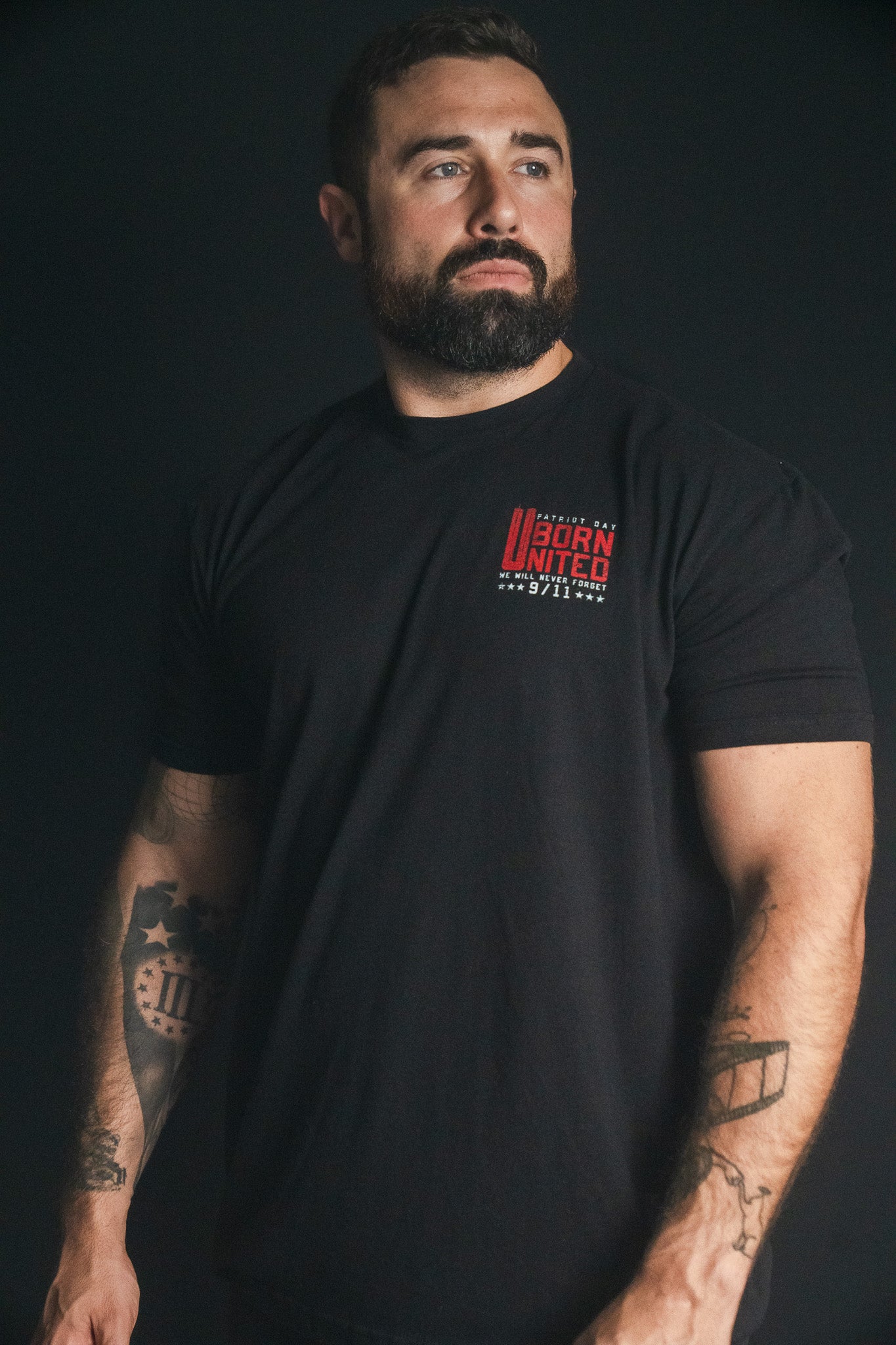A bearded man with tattoos wears the black “We Remember: 25th Anniversary” tee by Born United Private Label, standing against a dark background and looking to the side—a quiet tribute to honor and resilience.