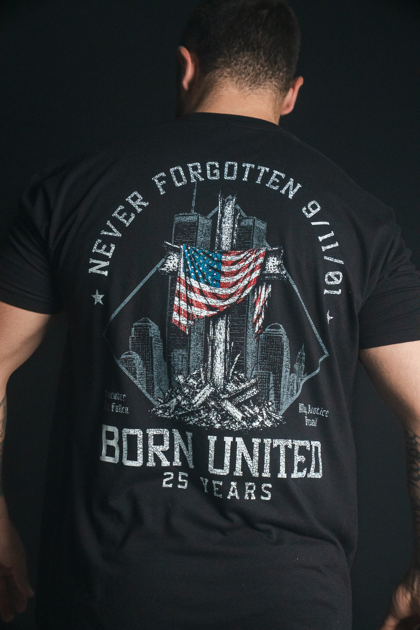 A person wearing the Born United Private Label “We Remember: 25th Anniversary” tee—a black remembrance shirt featuring a 9/11 memorial graphic, American flag, and the text “Never Forgotten 9/11/01” and “Born United 25 Years” on the back. This powerful tribute honors resilience and commemorates the legacy of September 11.