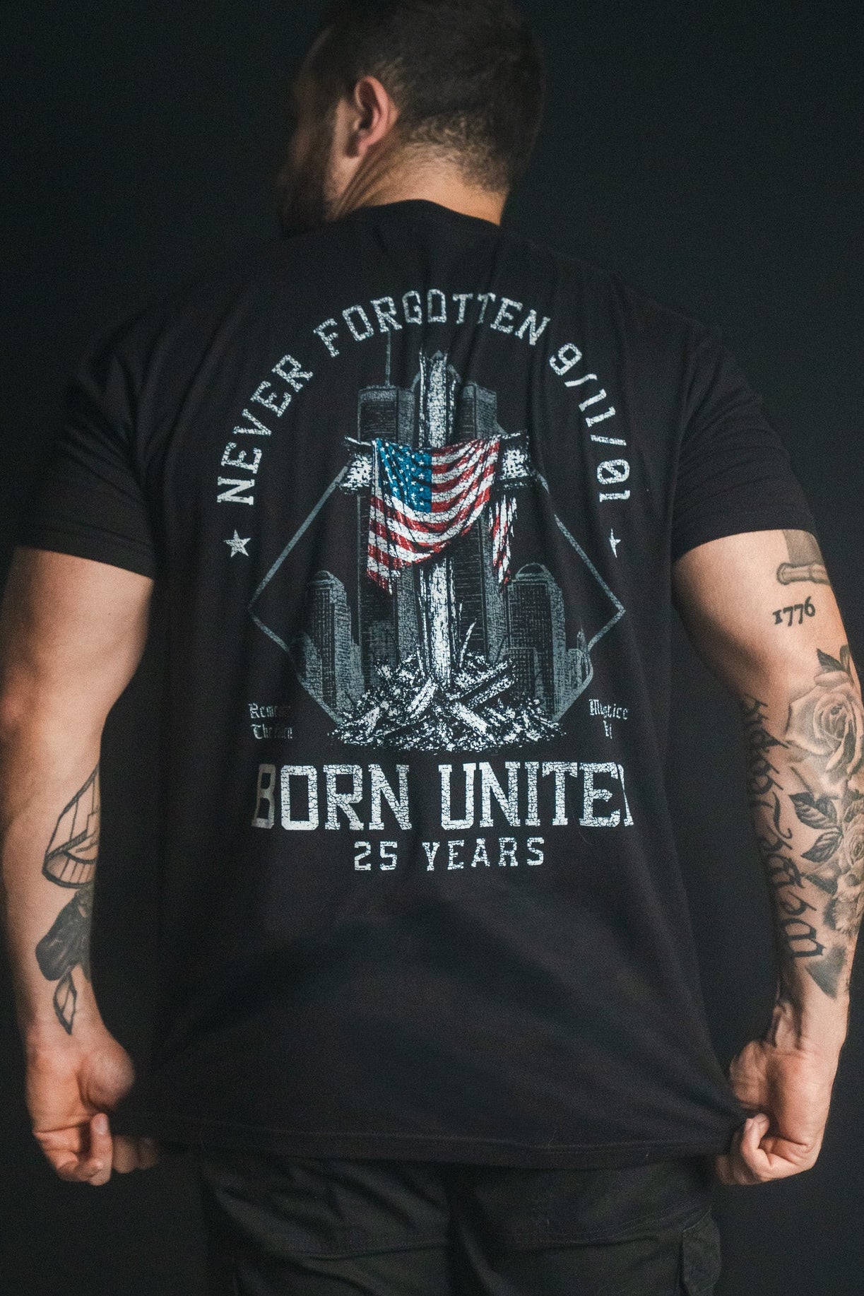 A person with tattoos wears the We Remember: 25th Anniversary tee by Born United Private Label—a black remembrance shirt featuring an American flag, twin towers, “Never Forgotten 9/11,” and “Born United 25 Years” in white text on the back, serving as a powerful September 11 memorial honoring resilience and unity.