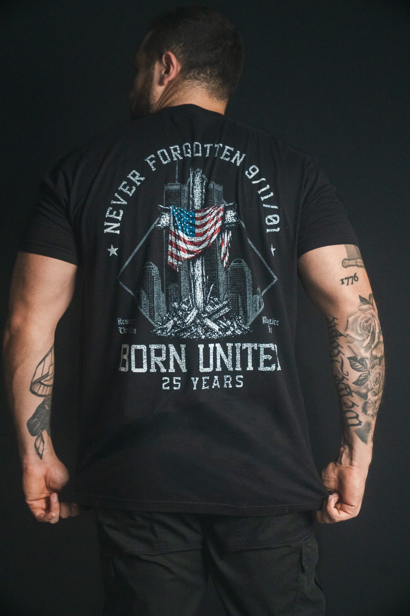 A person with tattoos wears the We Remember: 25th Anniversary tee by Born United Private Label—a black remembrance shirt featuring an American flag, twin towers, “Never Forgotten 9/11,” and “Born United 25 Years” in white text on the back, serving as a powerful September 11 memorial honoring resilience and unity.