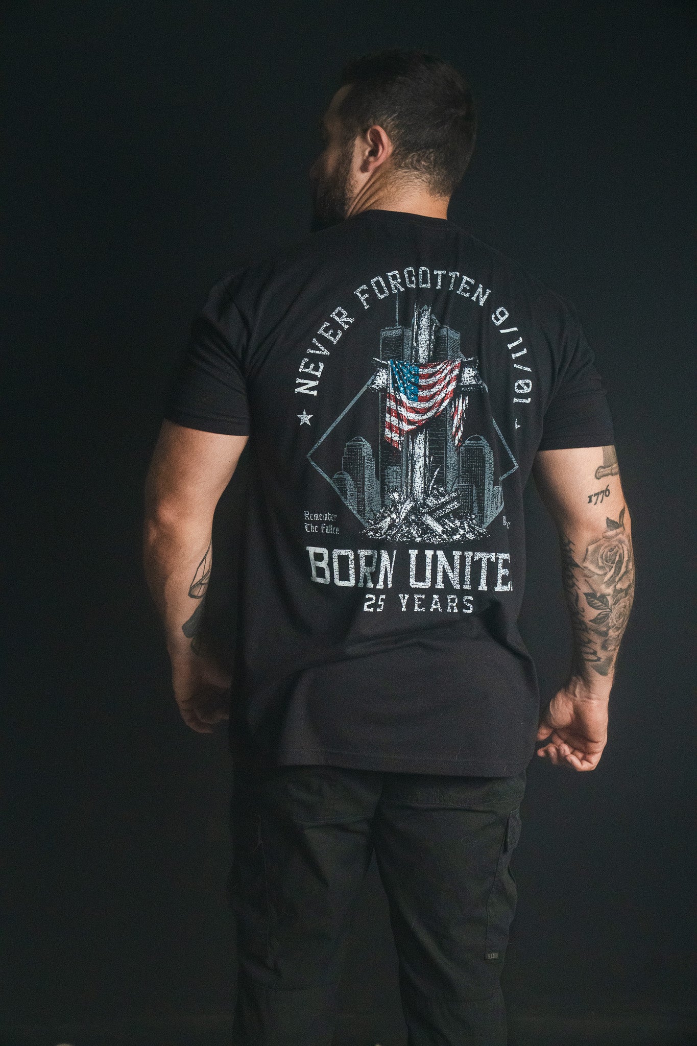 A man stands with his back to the camera, wearing a black We Remember: 25th Anniversary tee from Born United Private Label that features a September 11 memorial design and the text Never Forgotten 9/11—symbolizing honor and resilience.