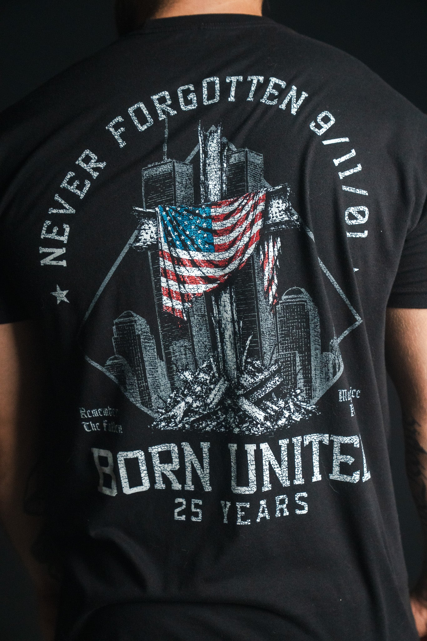 A person wearing the “We Remember: 25th Anniversary” black remembrance tee by Born United Private Label, featuring an illustration of the Twin Towers, an American flag, and the text Never Forgotten 9/11/01, Born United—a powerful September 11 memorial symbolizing honor and resilience.
