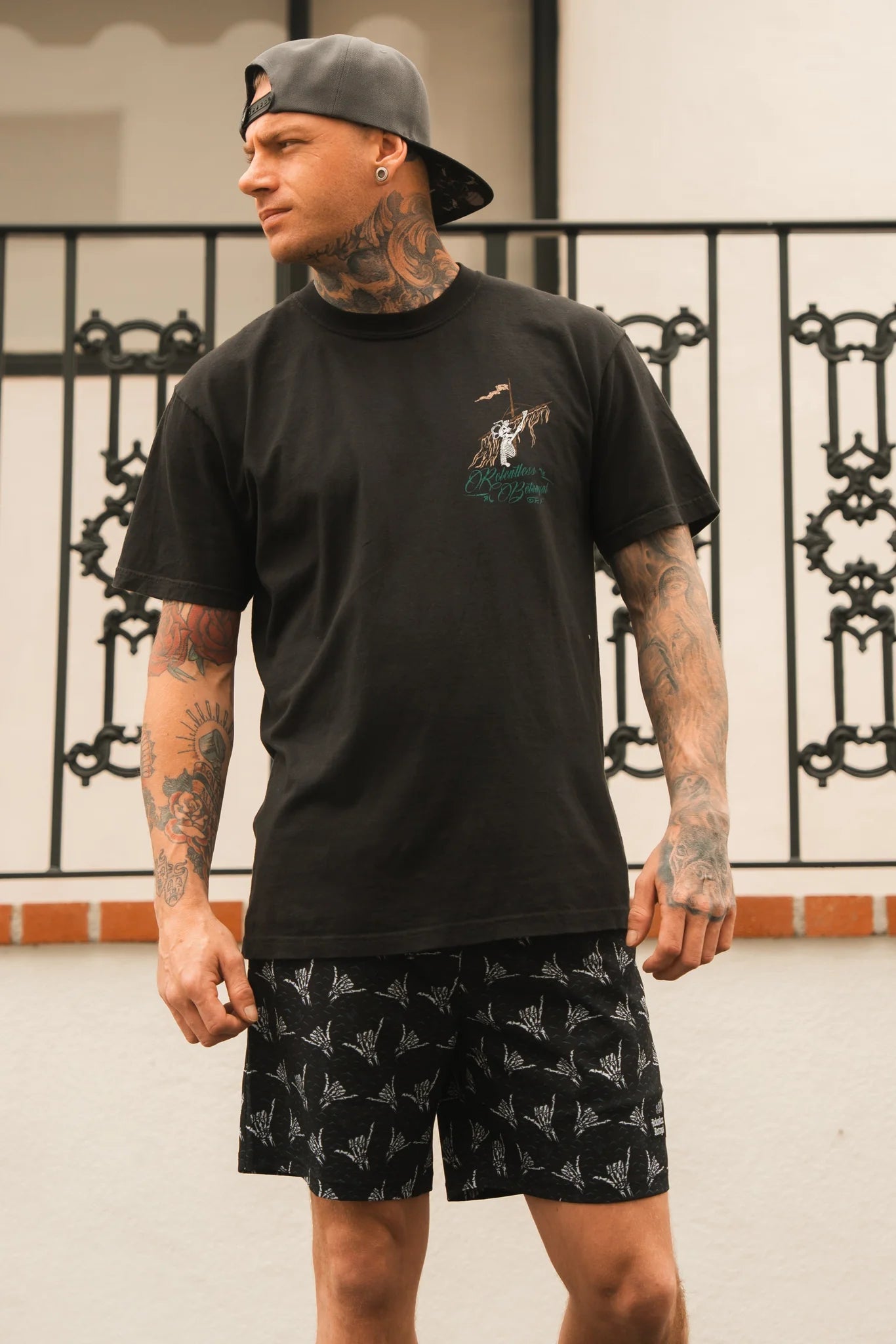 A tattooed man in a black graphic tee, backwards black cap, and Relentless Betrayal Mens Performance Athletic Shorts | 5.5- Sink or Swim stands outside by a decorative railing, looking to his left.
