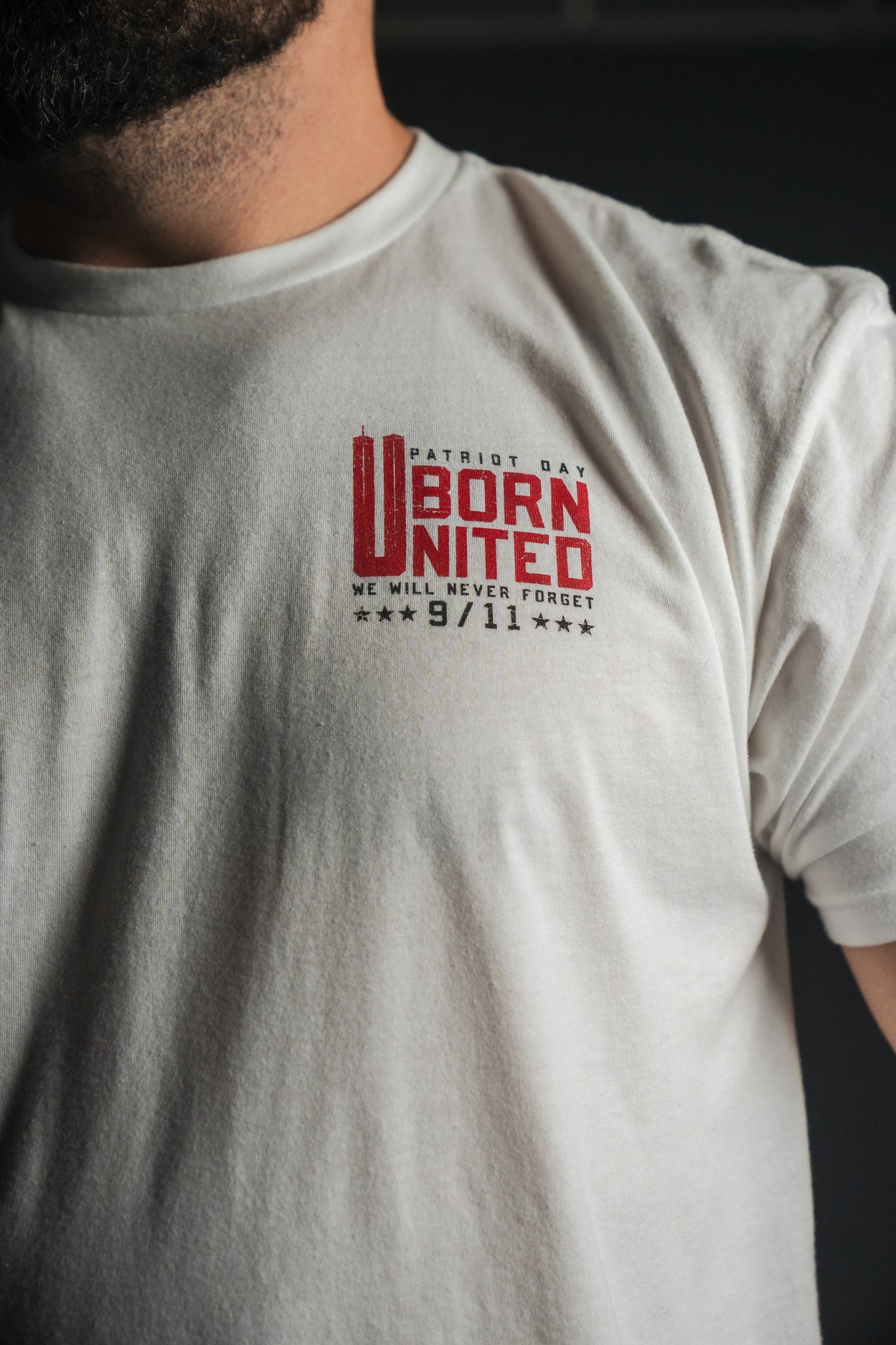 Wearing the “We Remember: 25th Anniversary - White” tee from Born United Private Label, featuring red “Patriot Day Born United We Will Never Forget 9/11” text on the left chest—a subtle yet powerful tribute to the September 11 memorial and a testament to honor and resilience.