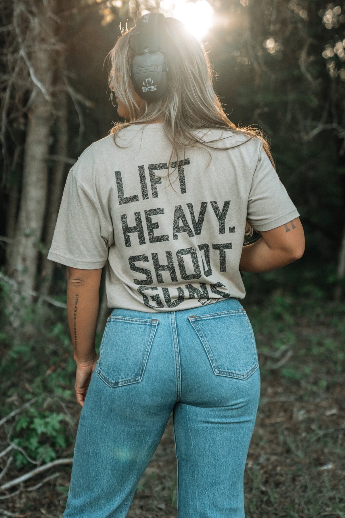 A person stands outdoors in jeans and a LHSG - Tan Tee by Tango Charlie Apparel—ideal for high-intensity fitness. Sunlight filters through the trees behind them.
