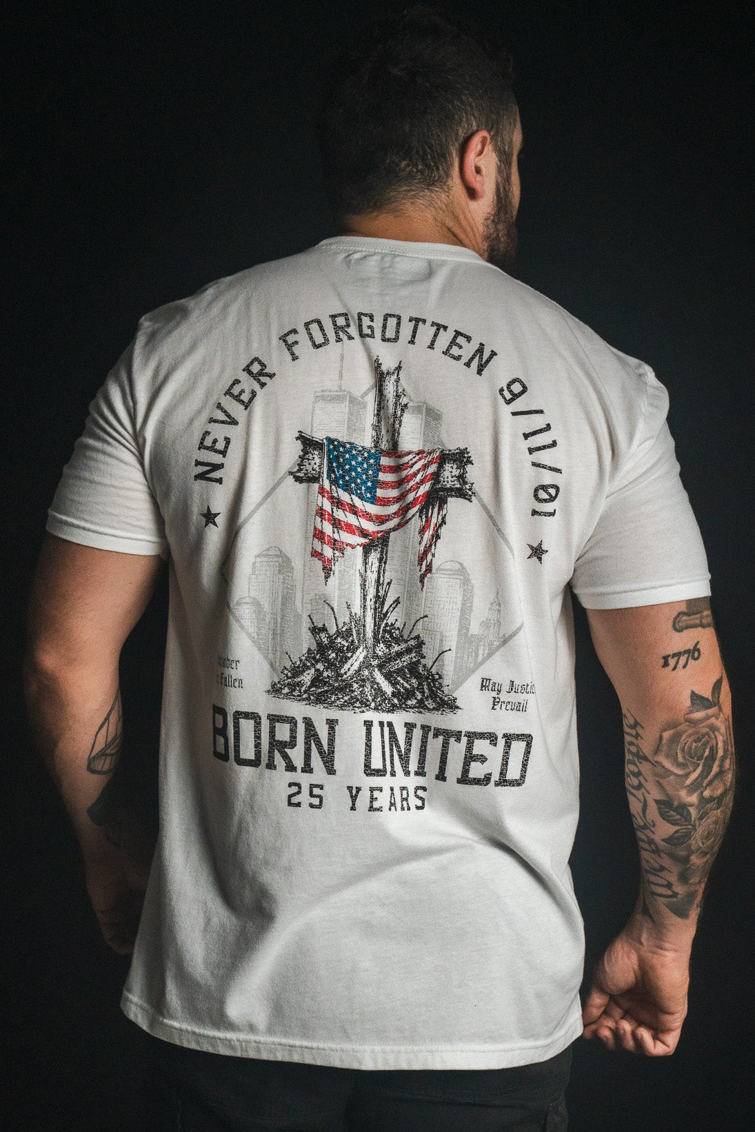 A man faces away wearing the “We Remember: 25th Anniversary - White” t-shirt by Born United Private Label, featuring an American flag, the text “Never Forgotten 9/11/01” and “Born United 25 Years,” and silhouettes of buildings—an inspiring September 11 memorial shirt honoring resilience and remembrance.