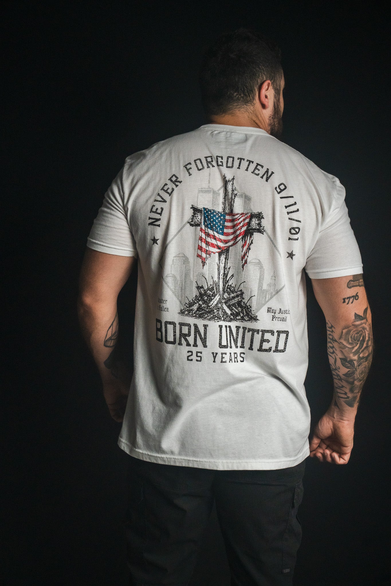 A man faces away wearing the “We Remember: 25th Anniversary - White” t-shirt by Born United Private Label, featuring an American flag, the text “Never Forgotten 9/11/01” and “Born United 25 Years,” and silhouettes of buildings—an inspiring September 11 memorial shirt honoring resilience and remembrance.