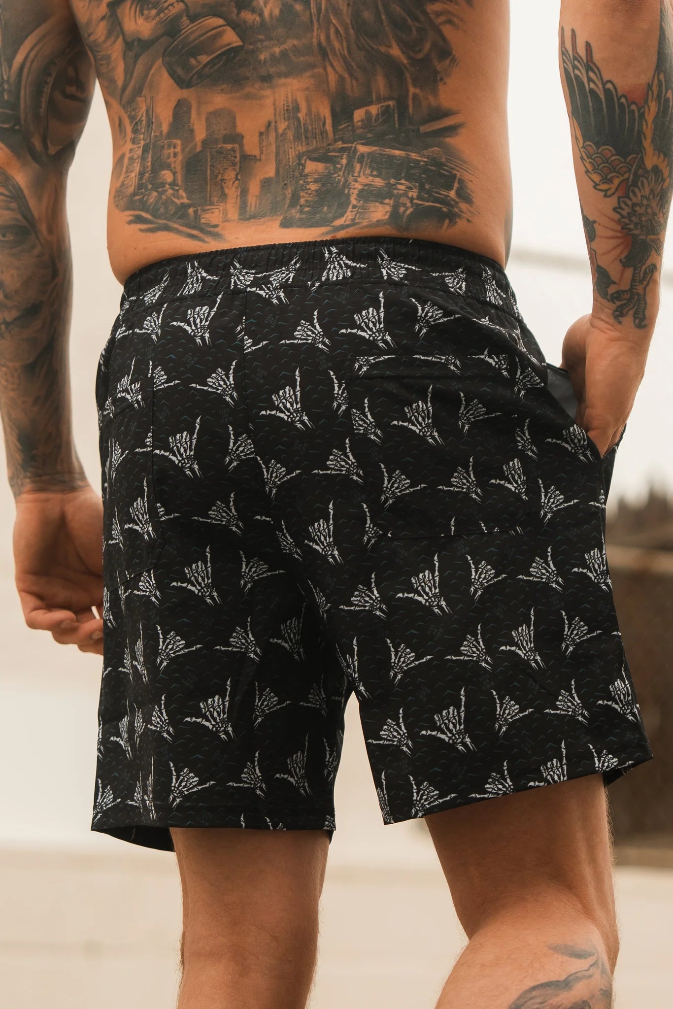 A tattooed man stands with his hand in his pocket, facing away from the camera, wearing Relentless Betrayal Mens Performance Athletic Shorts | 5.5–Sink or Swim, featuring a black and white botanical pattern.