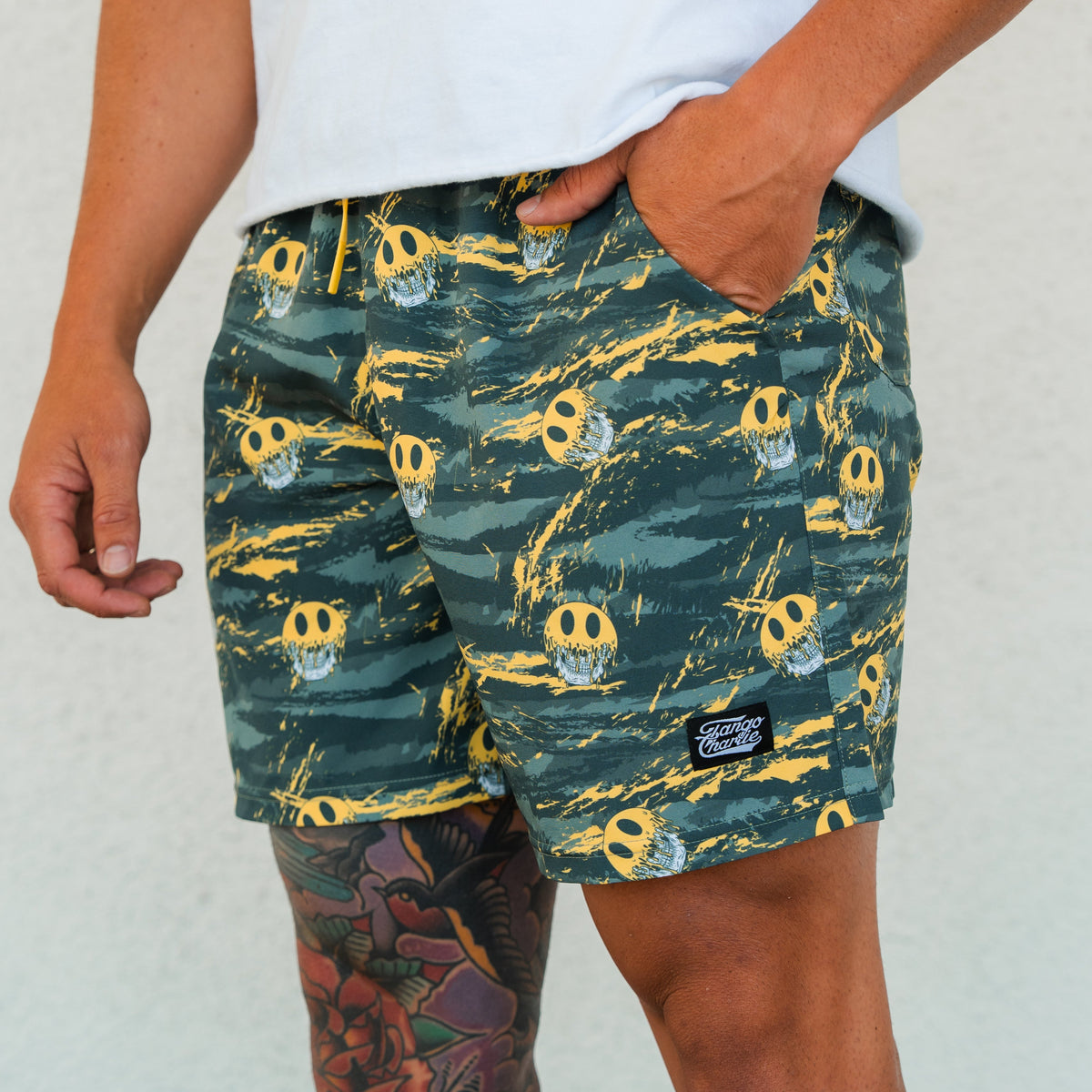 A person wearing Tango Charlie Apparel's Men's Performance Training Shorts | 5.5"- Savage in green and yellow with smiley faces and a black patch, paired with a white shirt. A colorful tattoo is visible on their upper thigh.