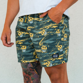 A person wearing Tango Charlie Apparel's Men's Performance Training Shorts | 5.5"- Savage in green and yellow with smiley faces and a black patch, paired with a white shirt. A colorful tattoo is visible on their upper thigh.
