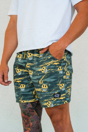 A person wearing a white T-shirt and Tango Charlie Apparel Men's Performance Training Shorts | 5.5"- Savage with a yellow skull pattern stands with one hand in their pocket, revealing part of a colorful tattoo on their thigh.