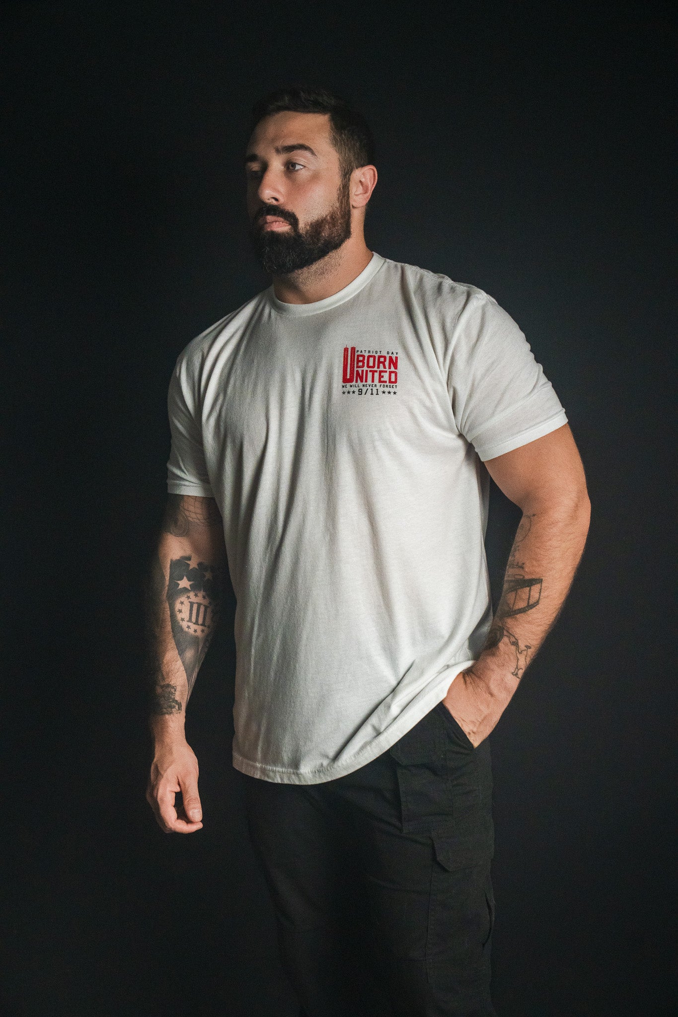 A bearded man stands against a dark background, wearing the We Remember: 25th Anniversary - White tee from Born United Private Label and black pants, with tattoos visible on both arms—a look that reflects honor and resilience.