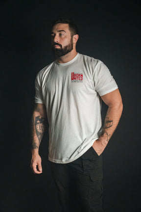 A bearded man stands against a dark background, wearing the We Remember: 25th Anniversary - White tee from Born United Private Label and black pants, with tattoos visible on both arms—a look that reflects honor and resilience.