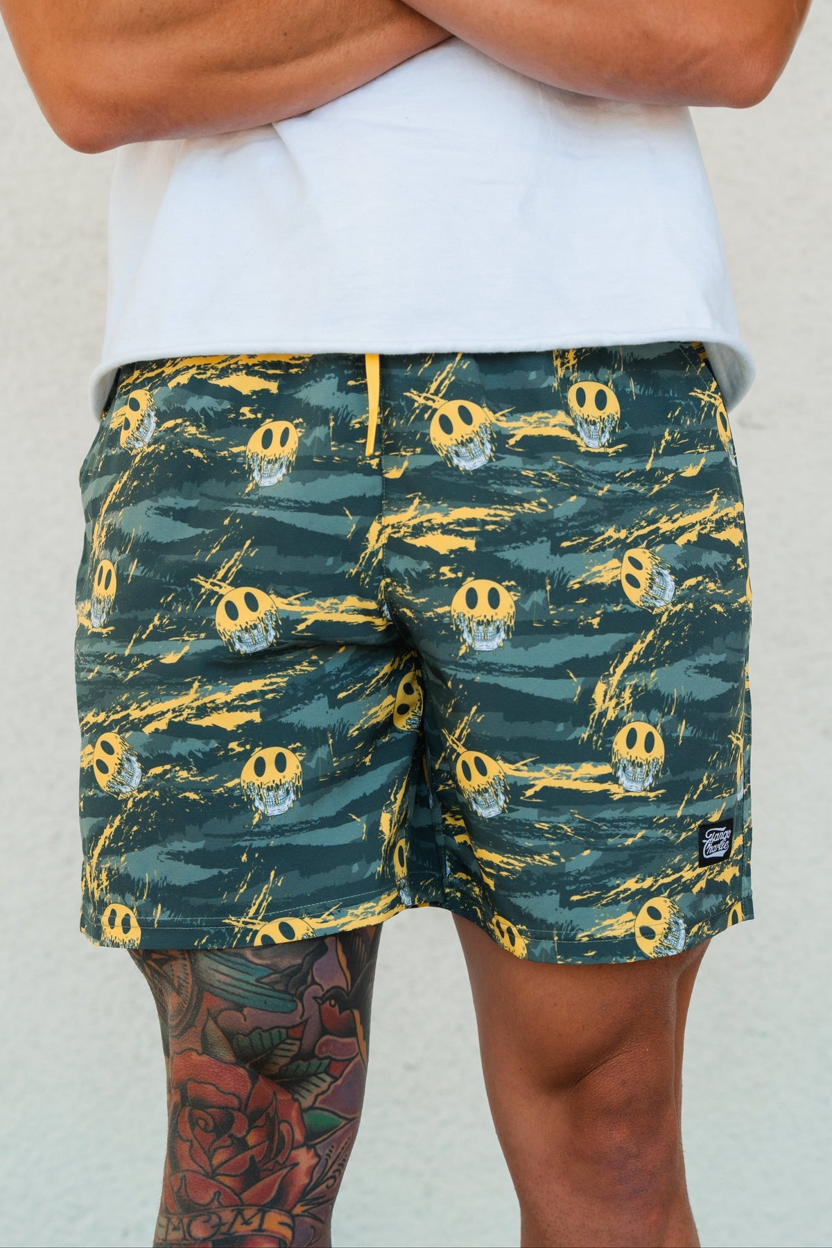 A person stands against a light wall, wearing Tango Charlie Apparel Men's Performance Training Shorts | 5.5"- Savage—green with yellow skulls—and a white shirt. A colorful tattoo is partly visible on their left thigh.