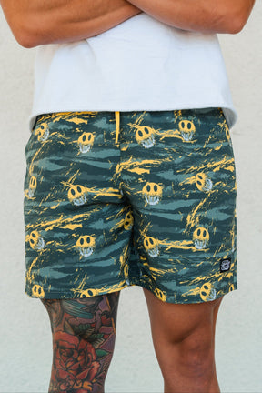 A person stands against a light wall, wearing Tango Charlie Apparel Men's Performance Training Shorts | 5.5"- Savage—green with yellow skulls—and a white shirt. A colorful tattoo is partly visible on their left thigh.