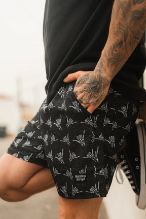A tattooed person stands outdoors wearing Relentless Betrayal Mens Performance Athletic Shorts | 5.5–Sink or Swim, featuring a white skeleton hand design and matching logo, with one hand in their pocket and black sneakers.
