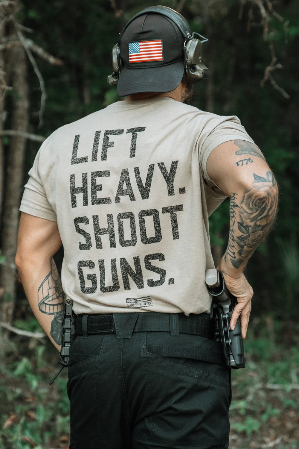 A person stands outdoors with their back to the camera, wearing the LHSG - Tan Tee by Tango Charlie Apparel—ear protection, tattoos, and a holstered pistol add to this high-intensity fitness gear look.