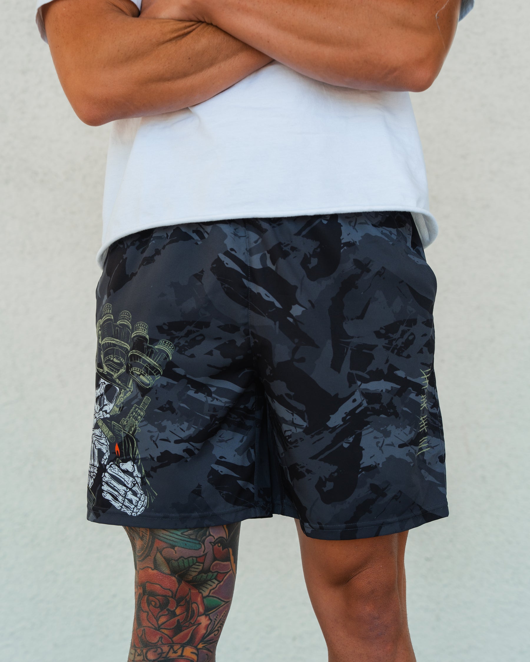 A person stands with arms crossed, wearing a white shirt and black Born United Private Label Mens Performance Hybrid Shorts 5.5 | Savage Ops, featuring a graphic design and tailored fit. A rose tattoo is visible on their leg against a plain light wall.