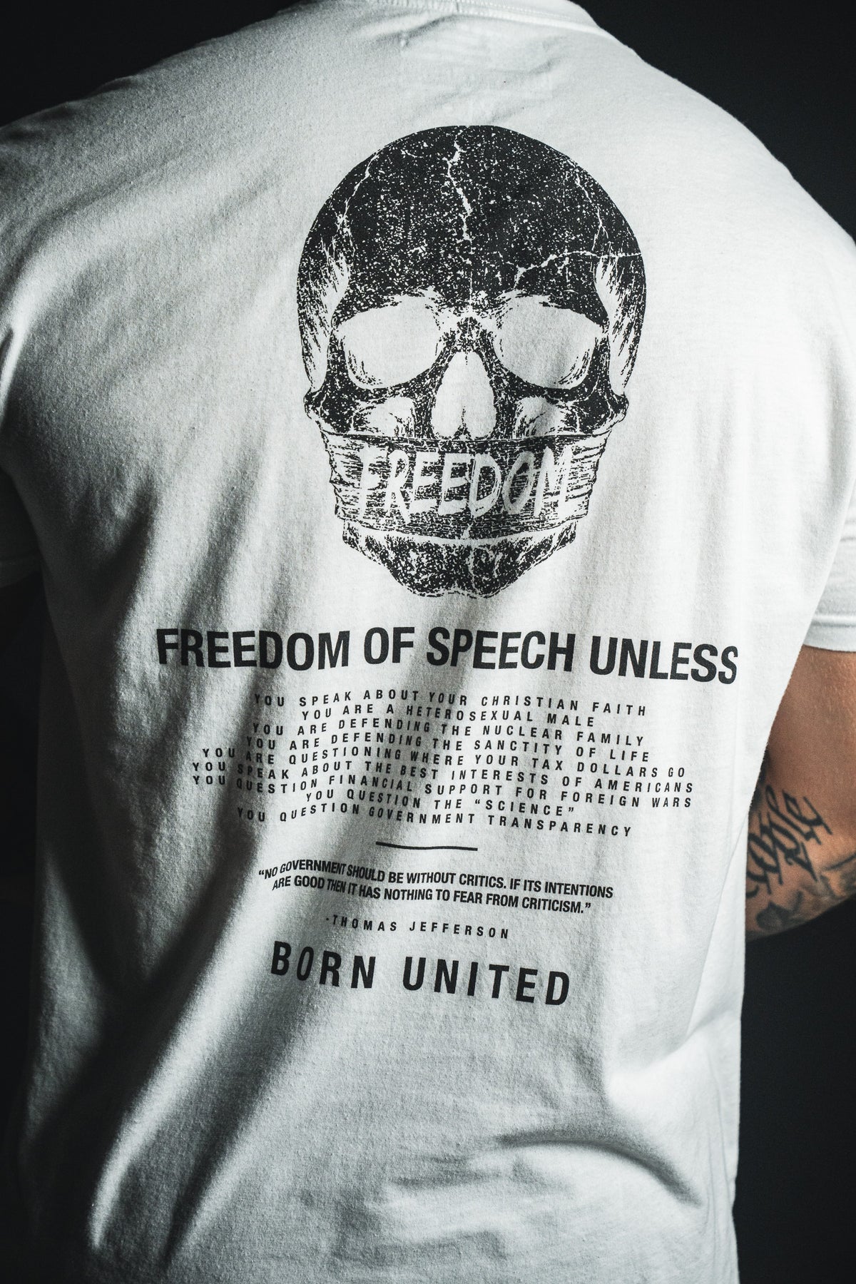 Wearing the Freedom of Speech - White t-shirt from Born United Private Label, this person sports a white tee displaying a black skull above the word “FREEDOM,” along with printed text about freedom of speech concluding in “BORN UNITED.” The design features subtle references to values such as the nuclear family and Christian faith. A tattoo is visible on their right arm.