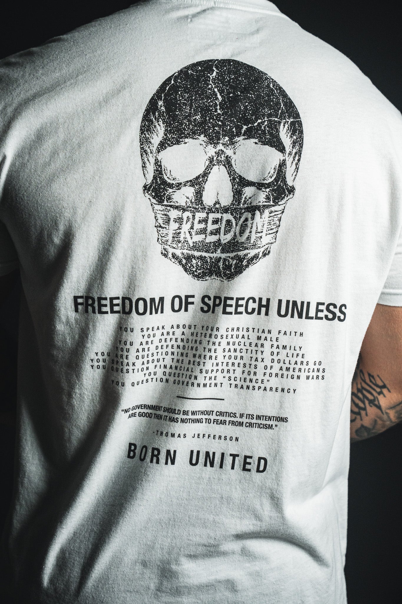 Wearing the Freedom of Speech - White t-shirt from Born United Private Label, this person sports a white tee displaying a black skull above the word “FREEDOM,” along with printed text about freedom of speech concluding in “BORN UNITED.” The design features subtle references to values such as the nuclear family and Christian faith. A tattoo is visible on their right arm.