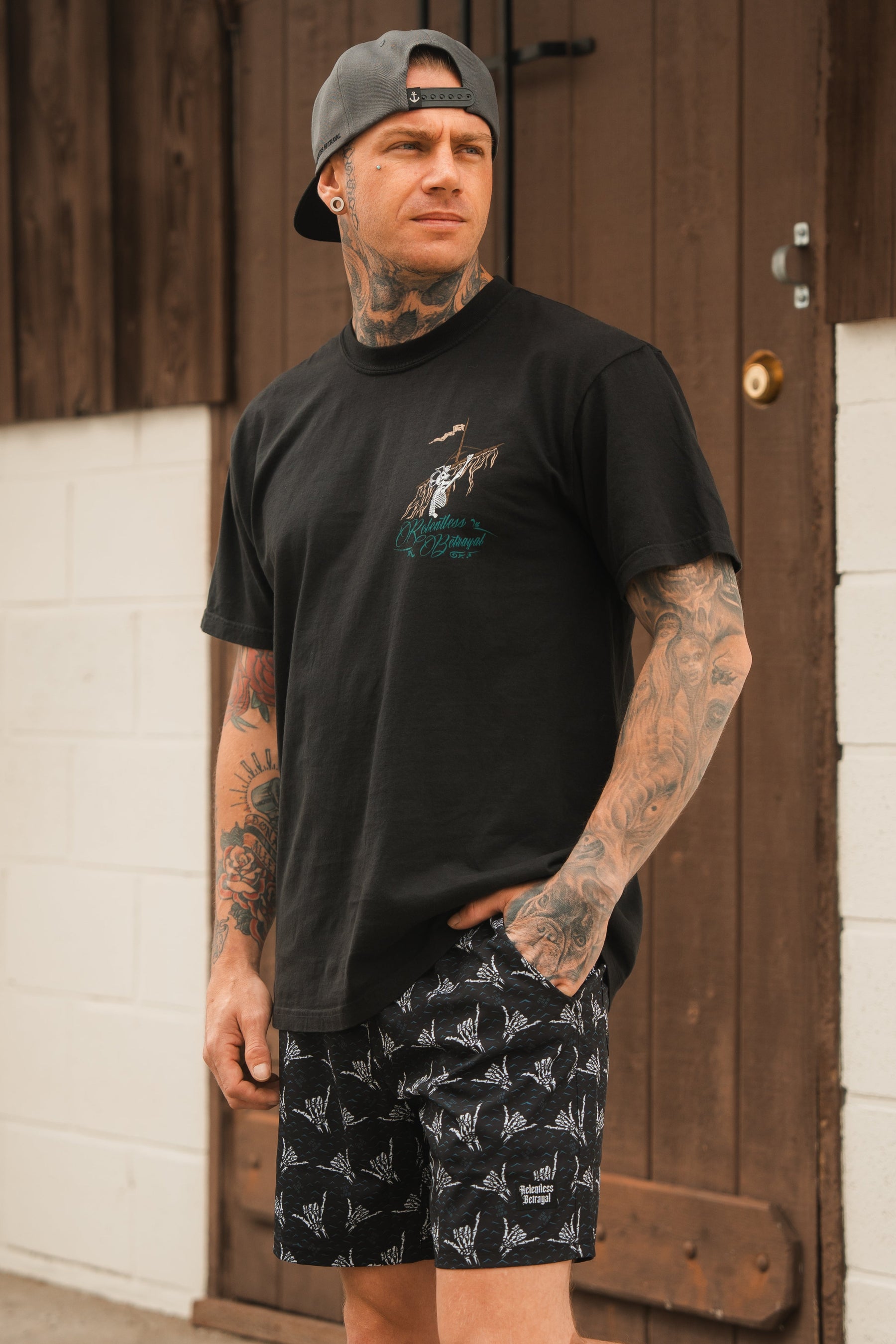 A tattooed man wearing the Relentless Betrayal Glorious Redemption Premium Tee—a 6th Anniversary Limited Edition made from premium jersey cotton—paired with patterned shorts and a backwards cap, stands in front of a brown wooden door, gazing to the side.