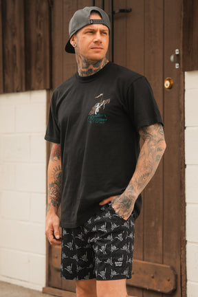 A tattooed man wearing the Relentless Betrayal Glorious Redemption Premium Tee—a 6th Anniversary Limited Edition made from premium jersey cotton—paired with patterned shorts and a backwards cap, stands in front of a brown wooden door, gazing to the side.