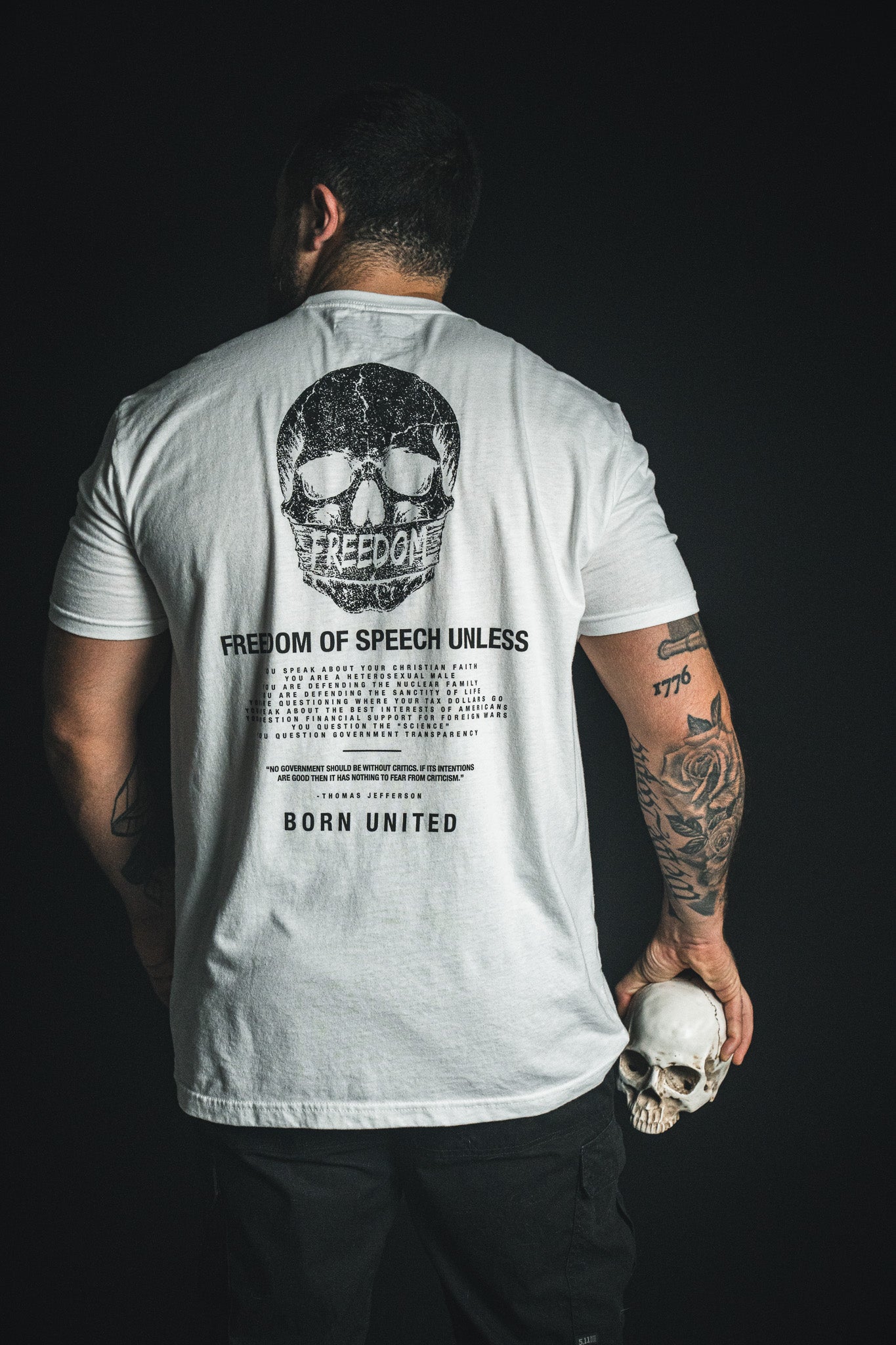 A tattooed man faces away, wearing the Born United Private Label “Freedom of Speech - White” t-shirt, which features a striking skull graphic and text. He holds a small skull in his left hand against a black background, making a bold statement.