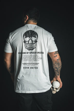 A tattooed man faces away, wearing the Born United Private Label “Freedom of Speech - White” t-shirt, which features a striking skull graphic and text. He holds a small skull in his left hand against a black background, making a bold statement.