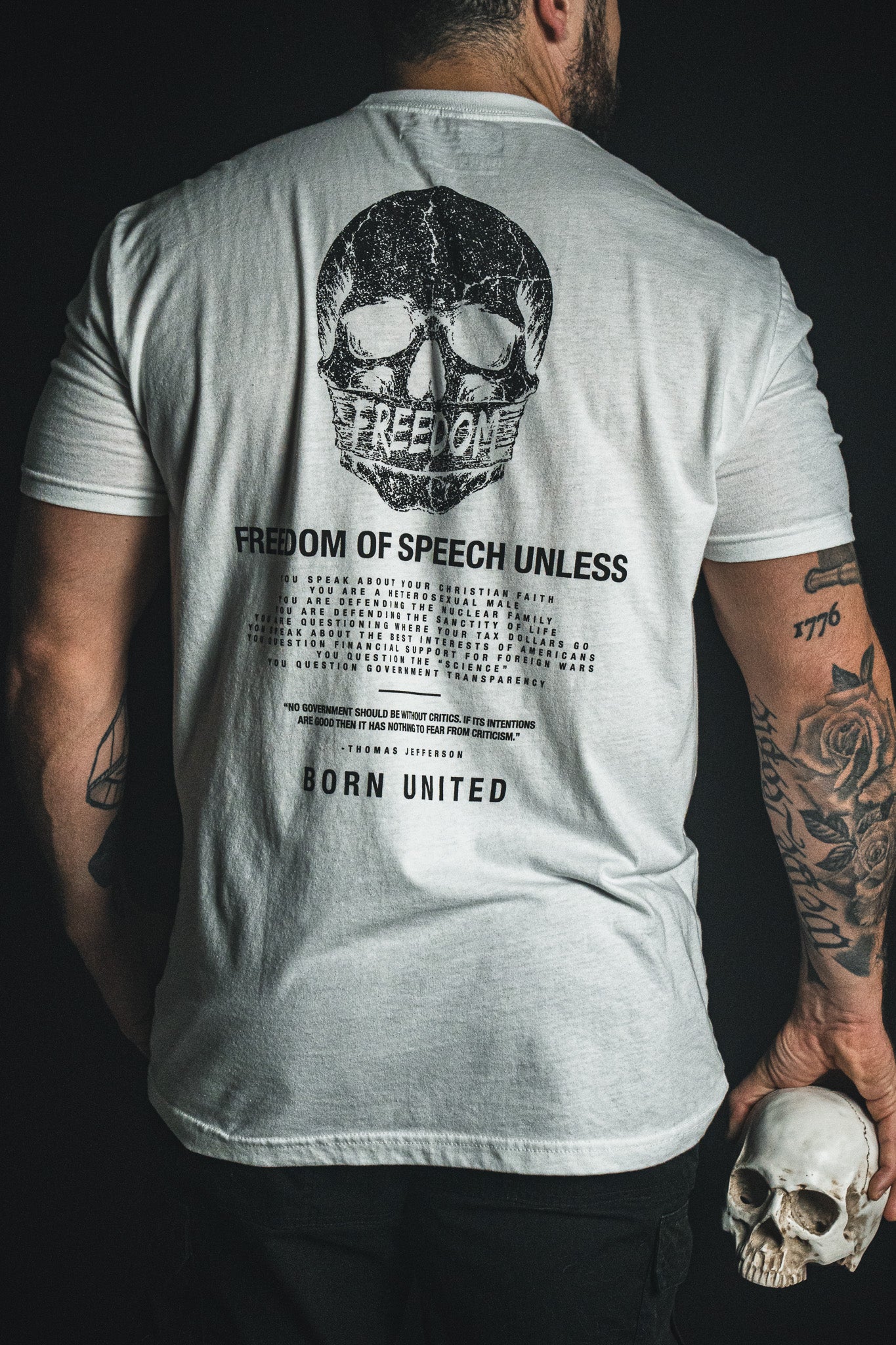 A tattooed individual holds a skull and wears the Born United Private Label’s “Freedom of Speech - White” T-shirt, featuring a large skull graphic and the words “FREEDOM OF SPEECH UNLESS” on the back, making a bold statement about freedom of speech and challenging conventional norms.