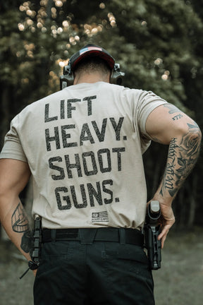 A muscular person, seen from behind, wears the LHSG - Tan Tee from Tango Charlie Apparel, with tattoos, hearing protection, and a holstered handgun visible against a blurred tree background.