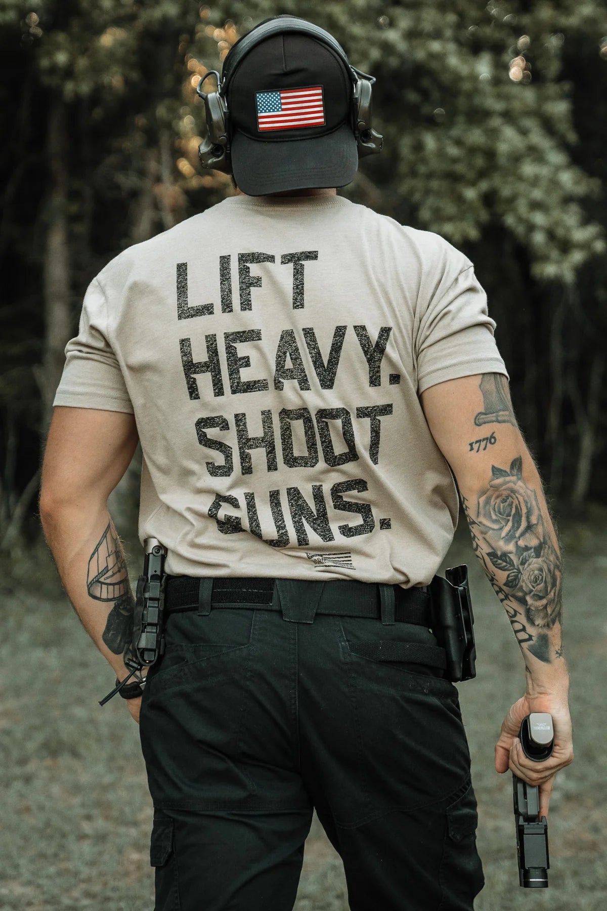Outdoors, a tattooed individual wears high-intensity fitness gear—a U.S. flag cap, ear protection, and the Tango Charlie Apparel LHSG - Tan Tee—holding a handgun with another holstered at their waist.