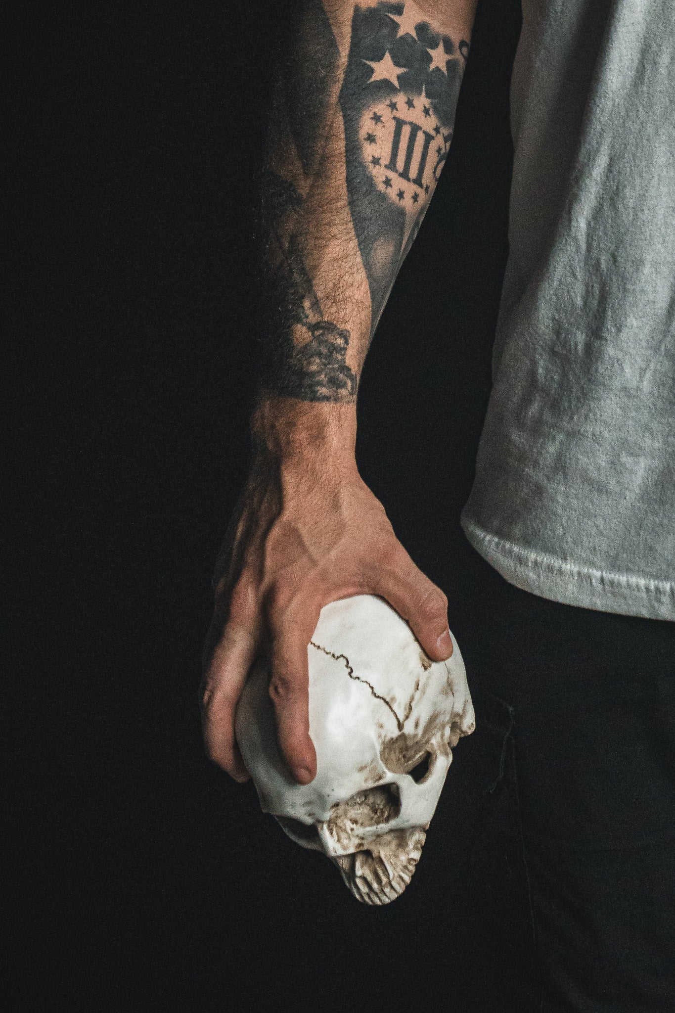 A tattooed individual wearing the “Freedom of Speech - White” shirt by Born United Private Label holds a white human skull in their right hand against a dark background, creating a striking image that prompts contemplation of themes like the Christian faith and profound questions surrounding life and mortality.