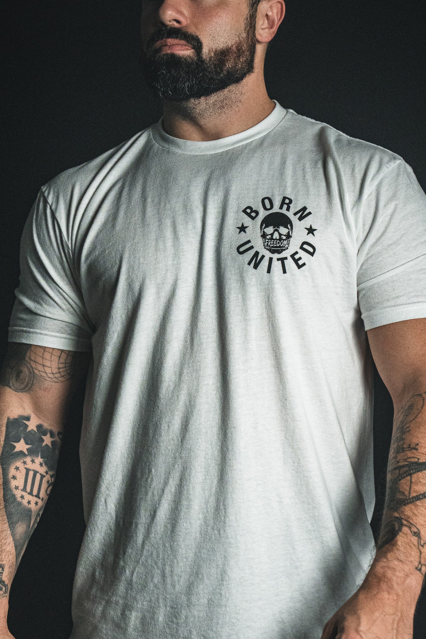 A bearded man stands against a dark background, displaying tattoos on both arms while wearing the “Freedom of Speech - White” t-shirt by Born United Private Label, embodying the values of Christian faith.