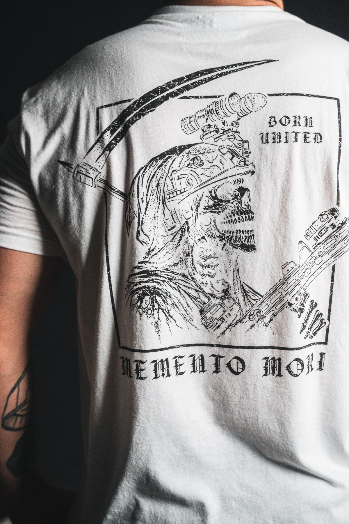 A person wearing the Memento Mori - White t-shirt from Born United Private Label, featuring a bold graphic of a grim reaper with a scythe and the text “Born United” and “Memento Mori” on the back—an eye-catching design that embraces philosophical reflections on mortality.